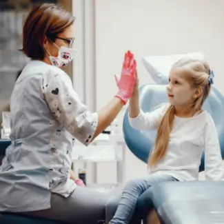 A dentist with a mask and gloves gives a high-five to a smiling young girl in a dental chair.