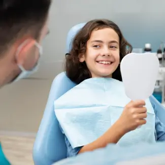 Boy in a dental chair smiling while holding a handheld mirror. A dentist in a mask, partially visible, engages with him.