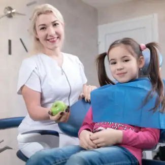 A smiling dentist in a white coat holds a green apple, standing next to a young girl with pigtails in a dental chair wearing a blue bib.