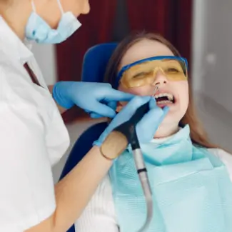 A dental professional in a mask and gloves works on a seated patient wearing protective glasses and a bib, holding dental tools.