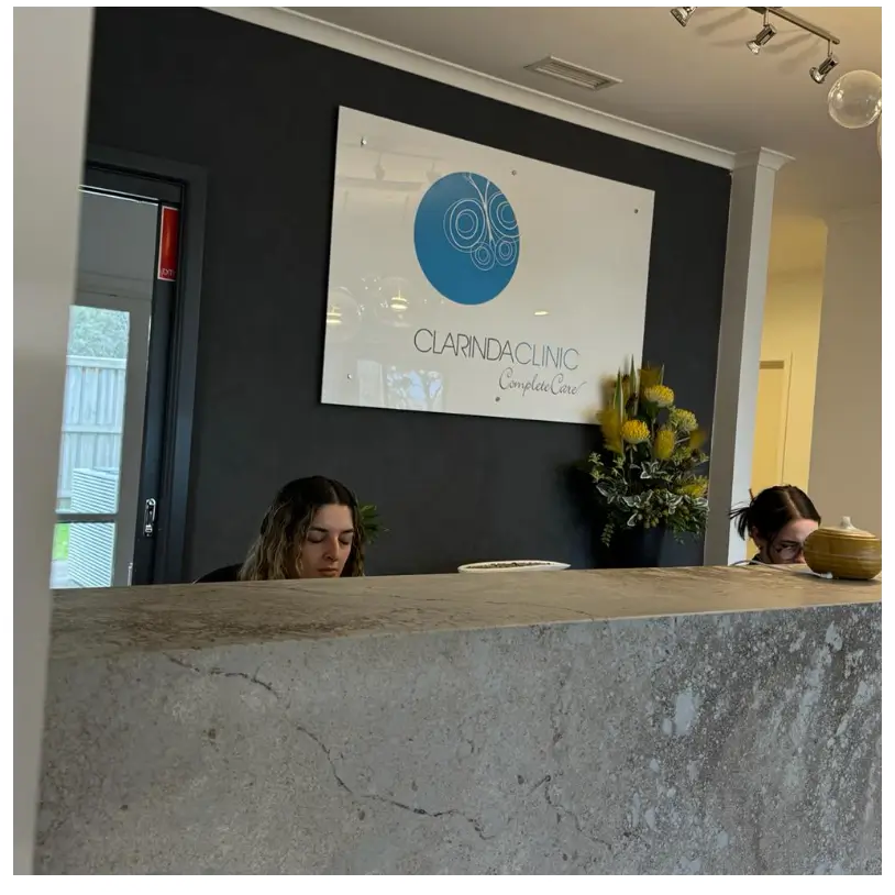 A calm reception area with a dark accent wall featuring the "Clarinda Clinic" sign. Two women are seated at the desk, focused on their tasks.