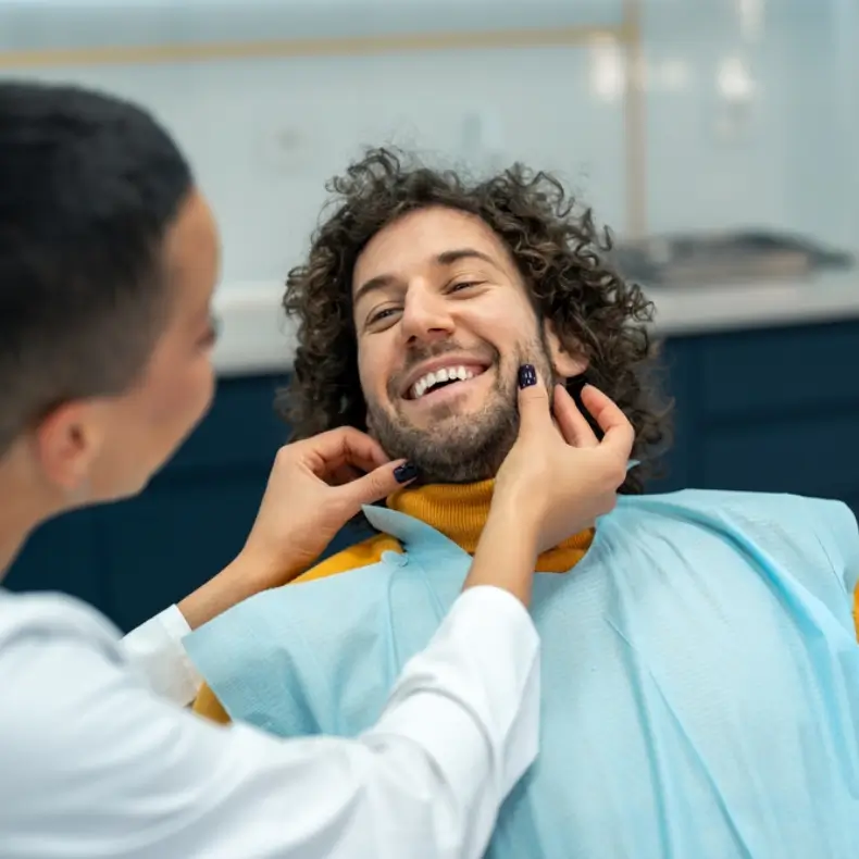A man with curly hair and a beard is smiling in a dental clinic, wearing a blue bib. A dentist is gently examining his face, creating a relaxed and cheerful atmosphere.