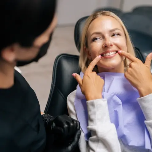 A woman in a dental chair smiles widely, pointing at her teeth with both hands. A dentist in black gloves looks on.