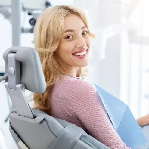 A woman with blonde hair smiles while seated in a dentist's chair, wearing a pink top and a blue dental bib. The setting is bright and clinical.