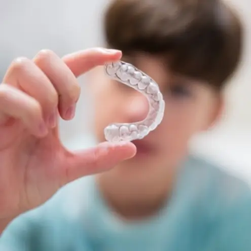 A person holds up a clear dental aligner in focus, with a blurred face in the background.
