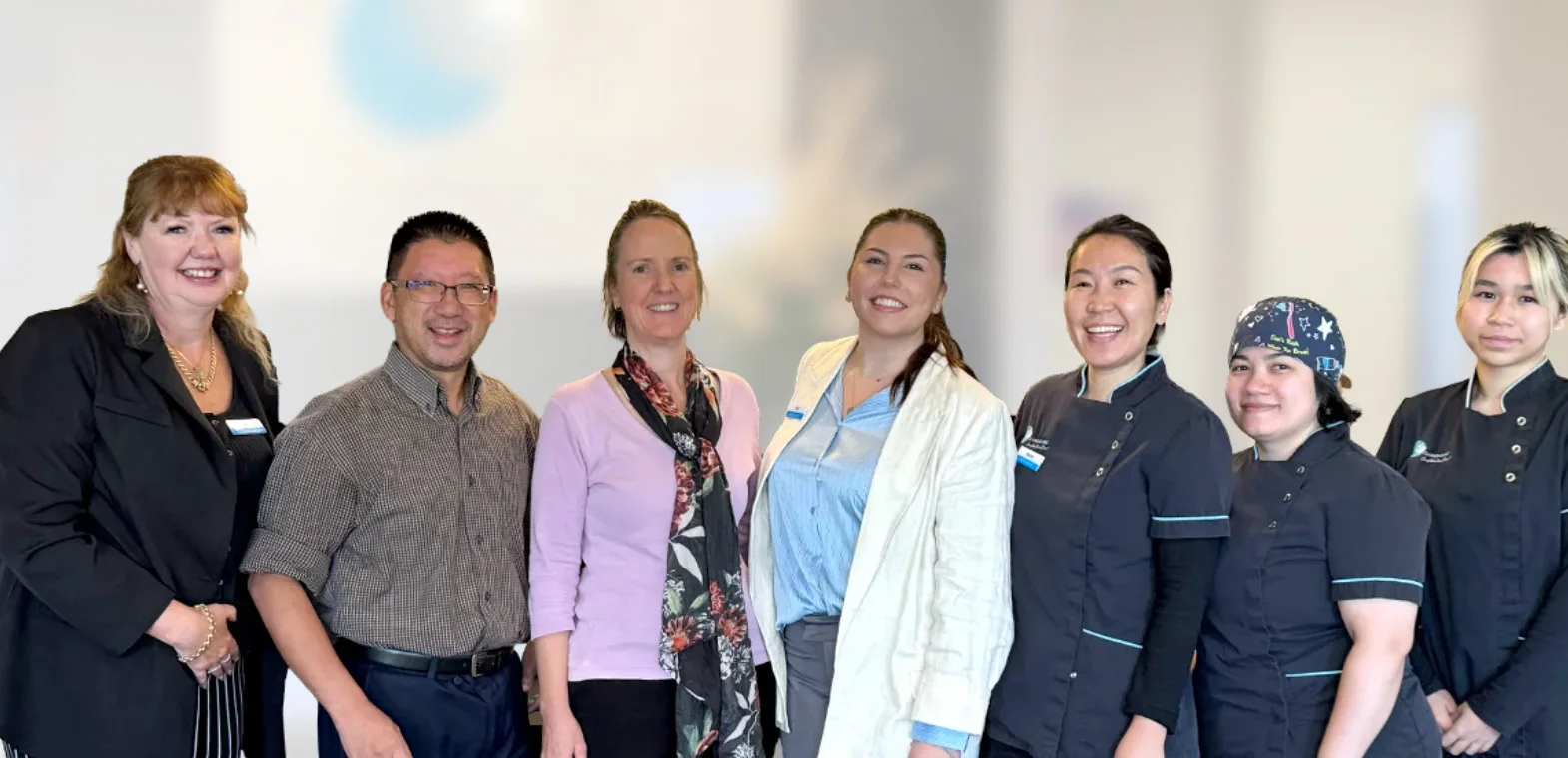 A dentist in blue scrubs and purple gloves gently adjusts a young girl's bib in a modern dental office. The girl, seated in an orange chair, smiles up at the dentist.