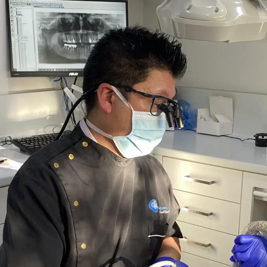 Dr Hong wearing magnifying glasses and mask examines patient with dental tools. X-ray on screen, bright and clinical setting.