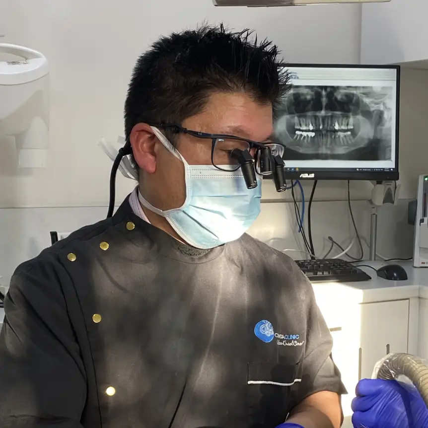 Dr Hong wearing a mask and magnifying glasses treats a patient lying down, using dental instruments. An X-ray is visible on a monitor. Bright, clinical setting.