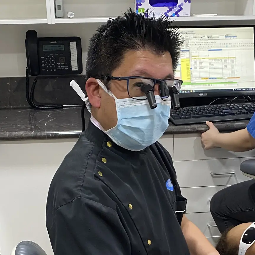 Dr Hong wearing a mask and gloves works on a patient lying in a chair, with an assistant at a computer in a modern dental office. The atmosphere is professional and clinical.