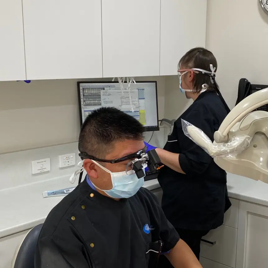 Dr Hong in a mask and gloves examines a patient under an overhead light in a clinic. Dr Snell works at a computer nearby.