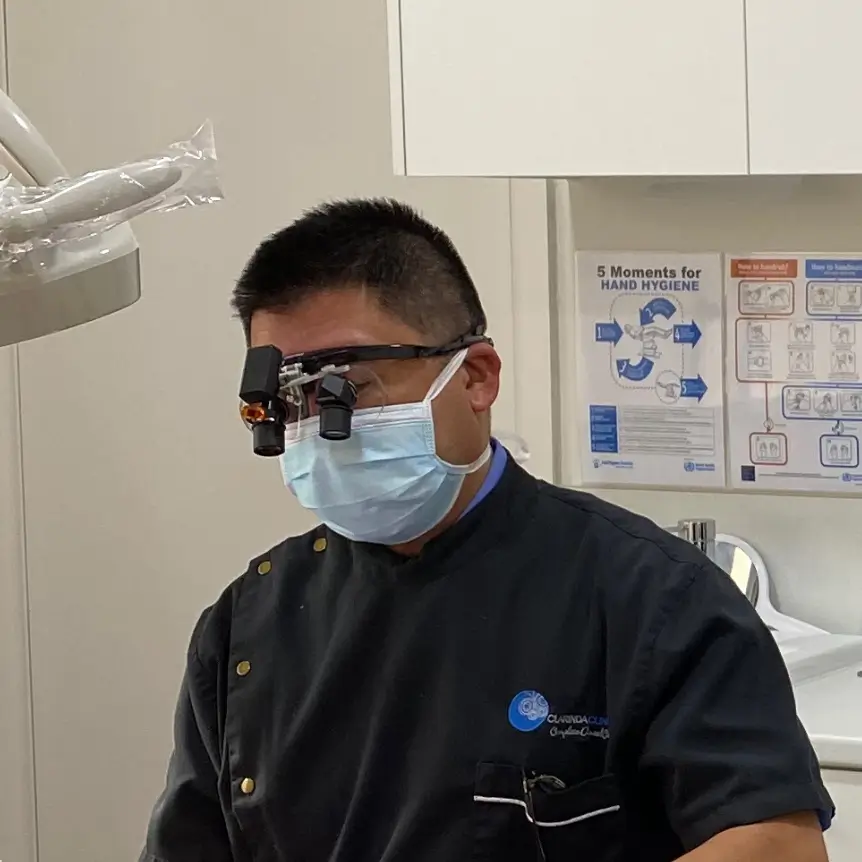 Dr Hong wearing magnifying glasses and mask works on a patient in a dental chair. An assistant stands by a computer in the modern, clean clinic.