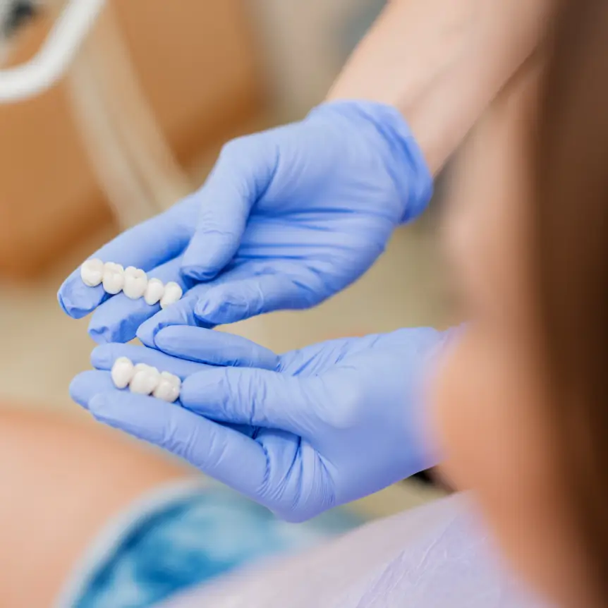 A dentist wearing blue gloves holds dental veneers, showing them to a patient. The image conveys a professional and clinical setting.