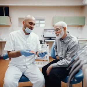 Dentist in white uniform and mask explains dental model to elderly male patient in gray shirt. They sit in a bright, modern dental office.