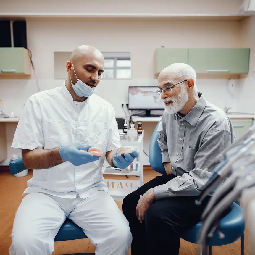 Dentist in white uniform and mask explains dental model to elderly male patient in gray shirt. They sit in a bright, modern dental office.