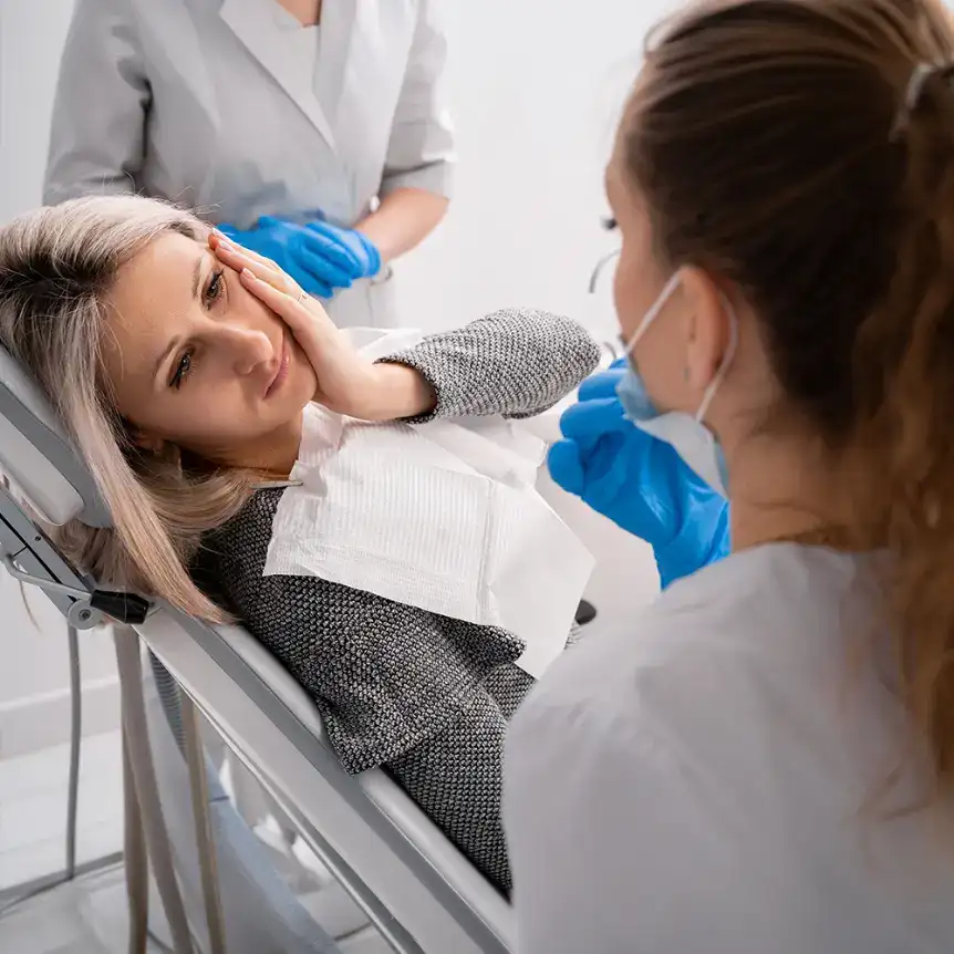 A woman in a dental chair holds her cheek in discomfort while a dentist in blue gloves and mask examines her. The setting is clinical and focused on dental care.