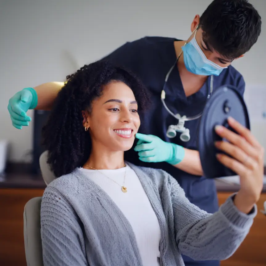 A woman with curly hair smiles while holding a mirror, sitting in a dental office. A dentist in scrubs and mask points, wearing gloves. The mood is positive.
