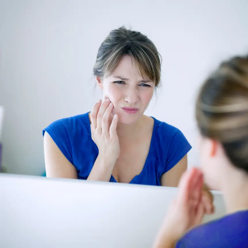 A woman in a blue shirt looks at her reflection in a mirror, touching her cheek with a pained expression, suggesting toothache or jaw pain.