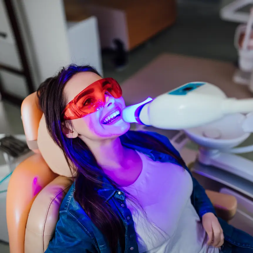 A woman in a dentist chair undergoes teeth whitening, wearing red protective glasses. A blue light shines on her teeth, creating a clinical, focused mood.