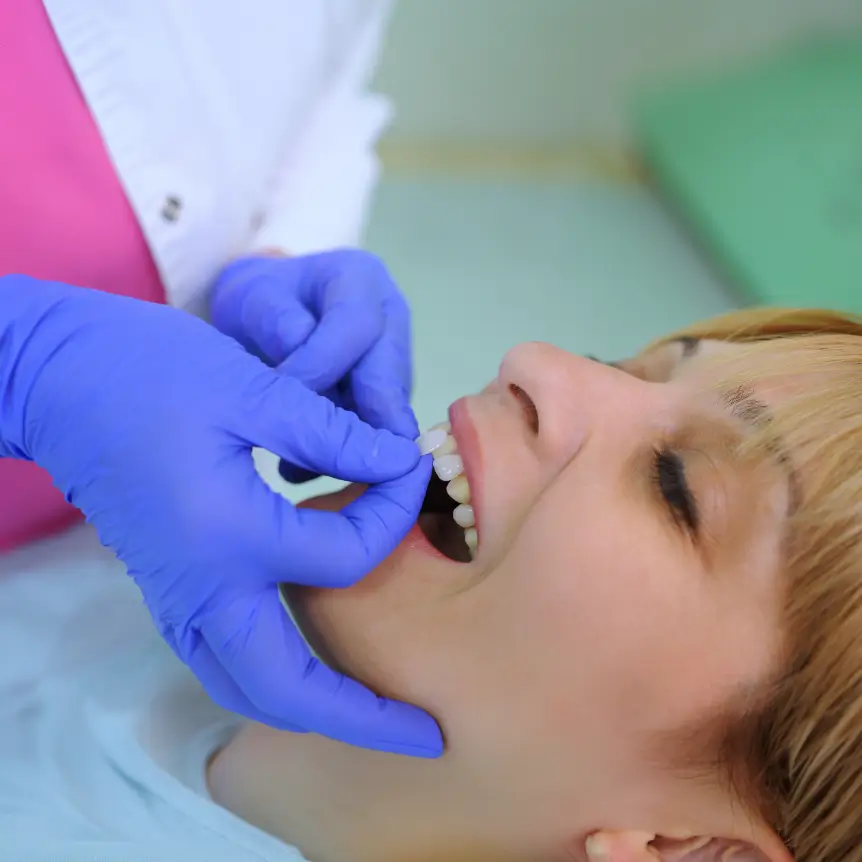 A dentist wearing blue gloves places a dental veneer on a smiling woman's tooth. The setting is clinical and professional, conveying a sense of care.