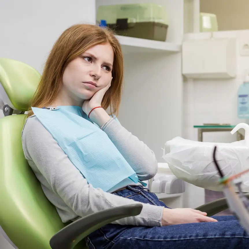 A woman sits in a dentist's chair, looking bored and resting her head on her hand. She wears a dental bib, with equipment visible in the background.