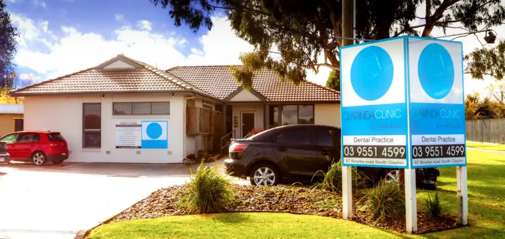 Single-story of Clarinda Clinic Exterior 2011 with grey roof, blue signage, and large windows. Front parking area, surrounded by trees and greenery, creating a welcoming atmosphere.