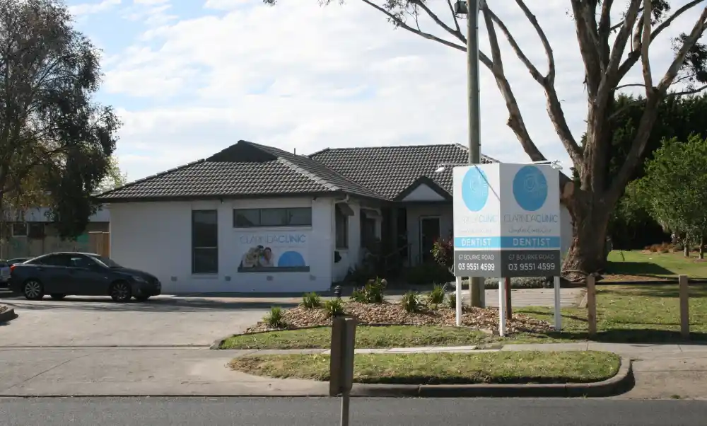 Single-story of Clarinda Clinic Exterior 2017 with grey roof, blue signage, and large windows. Front parking area, surrounded by trees and greenery, creating a welcoming atmosphere.
