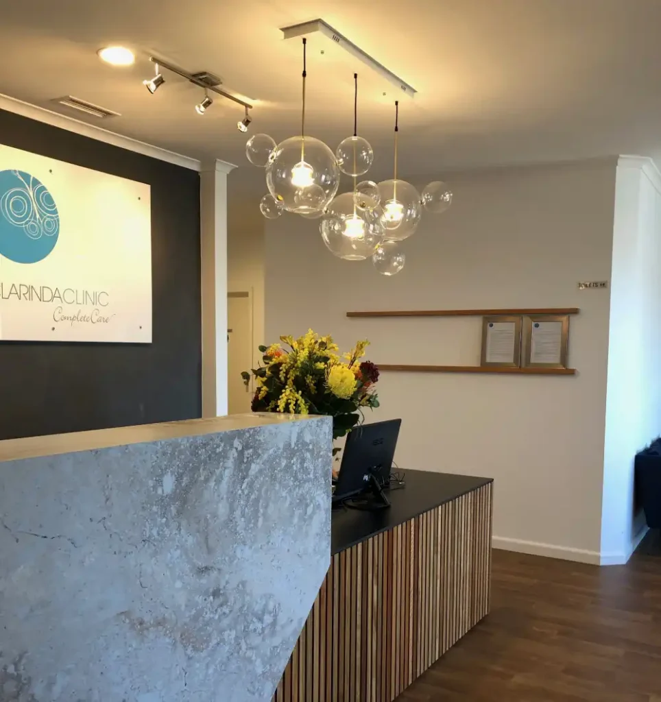 Modern Clarinda Clinic reception with a sleek concrete desk, pendant globe lights, wood accents, a vibrant flower arrangement, and a logo on the dark wall.