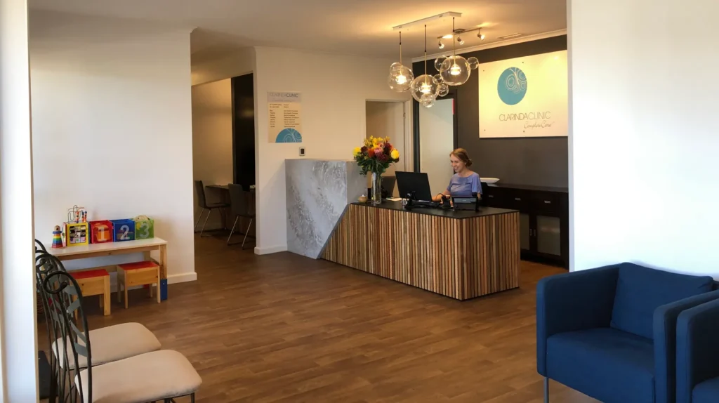 A welcoming Clarinda Clinic reception area with wooden floors, a receptionist at a modern desk, and decorative hanging lights. A play area with toys and seating are visible, conveying a warm, friendly atmosphere.
