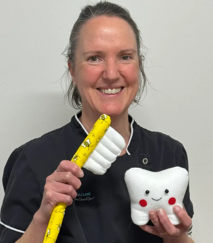 Smiling woman in a dental uniform holds a plush toothbrush and a cute, smiling tooth toy. The scene conveys a friendly and playful dental theme.