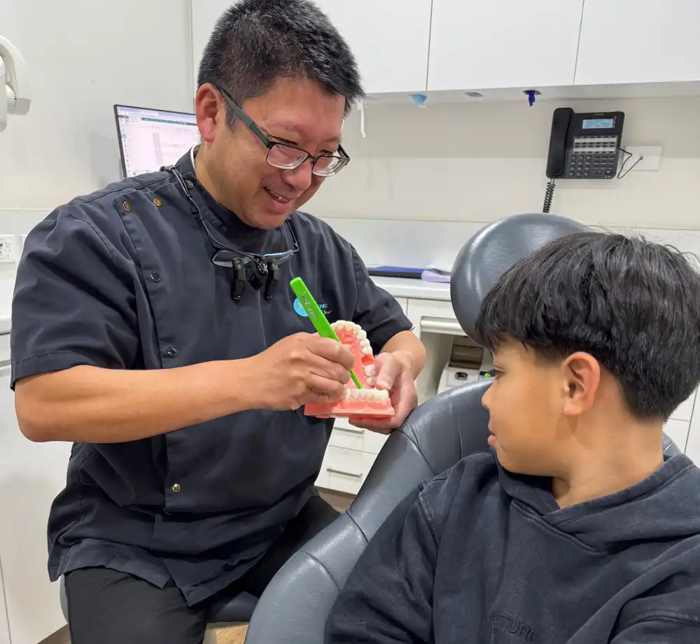 Dr Hong explaining dental care to a boy using a model of teeth. The dentist smiles while pointing at the model, creating a friendly atmosphere.