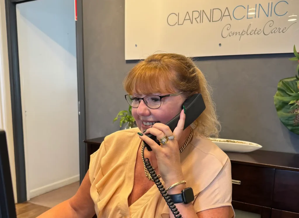 A woman with glasses, smiling and on the phone, sits at a desk with a keyboard. Behind her, a sign reads "Clarinda Clinic Complete Care."