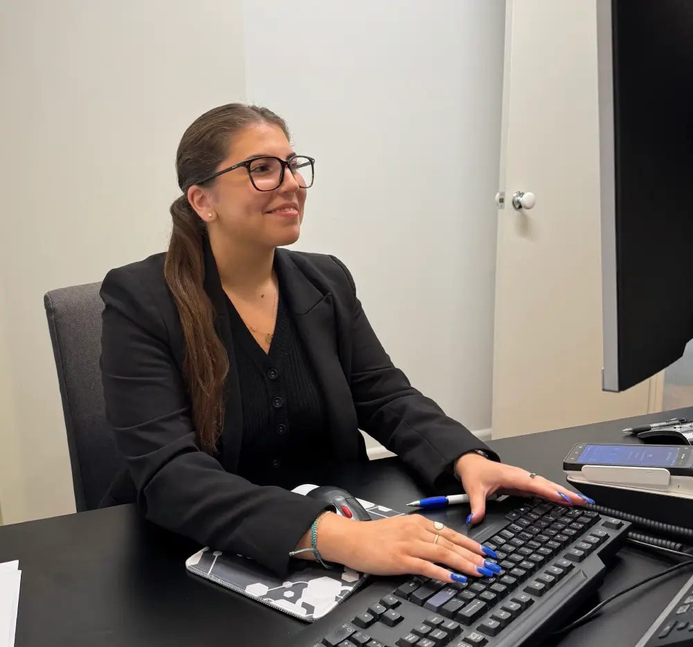 A woman in glasses and a black suit smiles while working at a computer. She uses a keyboard, with a notepad, pen, and smartphone nearby.
