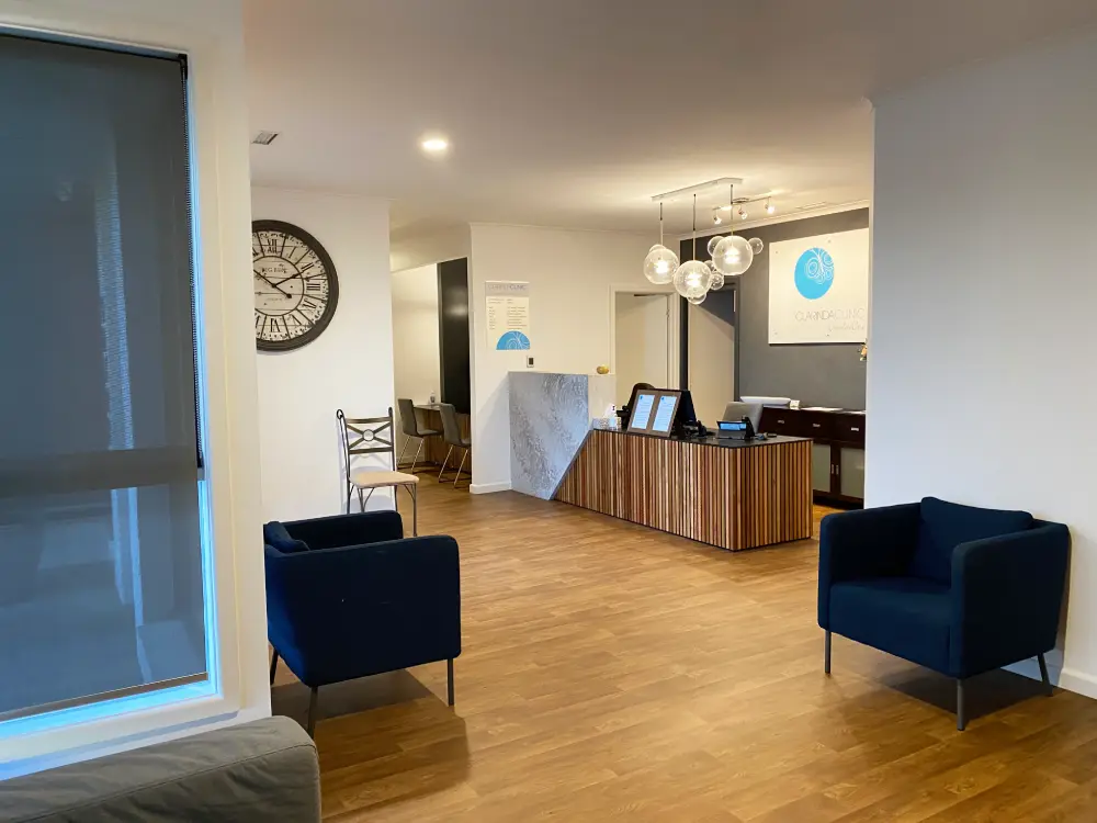 Clarinda Clinic a modern waiting room with wooden flooring, blue armchairs, and a large clock. A reception desk with globe lights and signage is visible in the background.