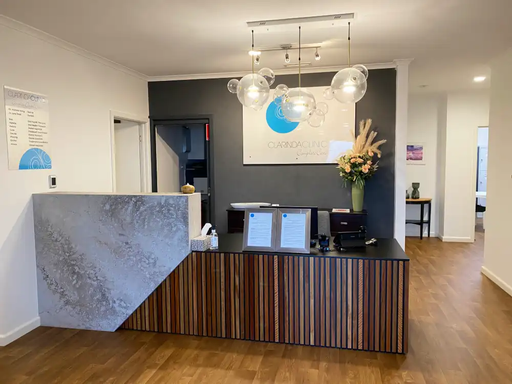 Reception area of Clarinda Clinic with a modern wooden desk, decorative pendant lights, and a flower arrangement. A sign with a blue logo is on the gray wall.