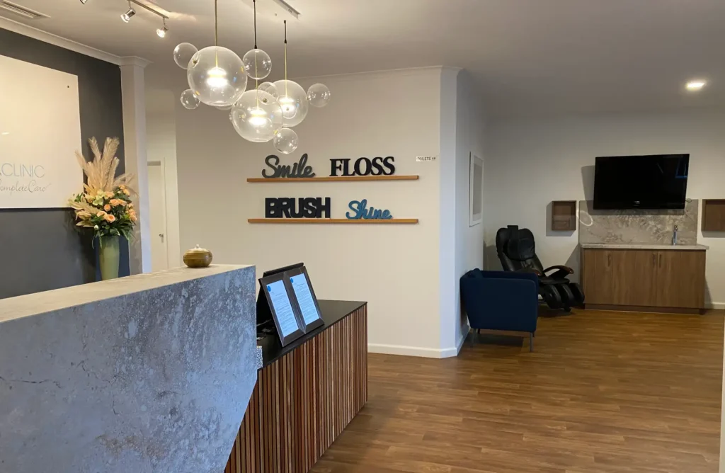 Reception area of a dental clinic with a modern decor. Wall features words "Smile," "Floss," "Brush," "Shine." Cozy seating and a TV nearby.