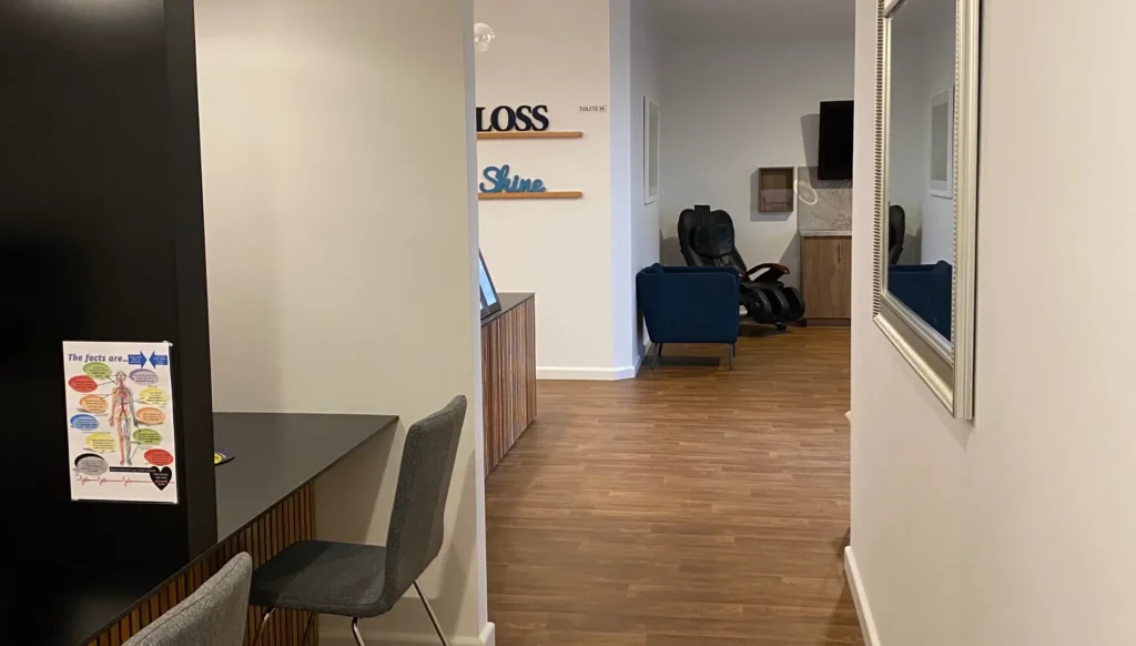Modern office interior with light wood flooring and a sleek design. Chairs face a black countertop with anatomical poster. Cozy seating area visible in the background.