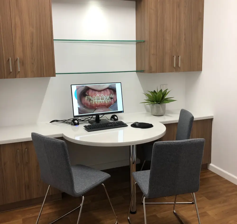 Dental consultation room with a round white table, two gray chairs, and wood cabinets. A computer displays a dental image. A potted plant adds a fresh touch.