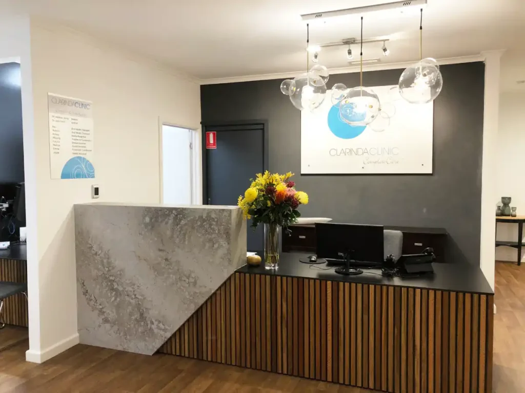 Clarinda Clinic Modern clinic reception with wood-accented desk, grey walls, and bright chandelier. A vase of yellow and red flowers adds a welcoming touch.